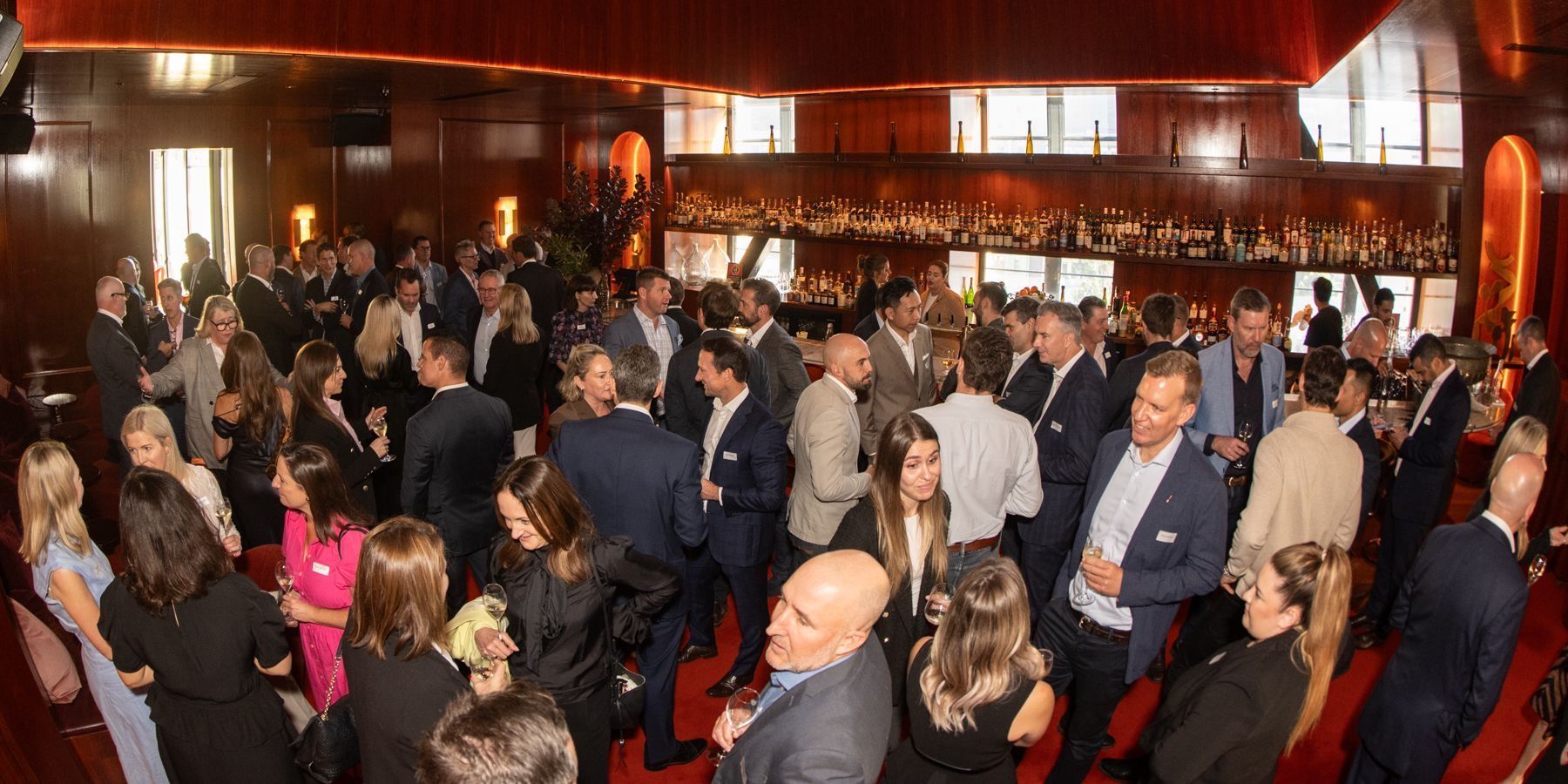A large group of people in business formal attire socializing and networking at an evening event in a wood-paneled room.