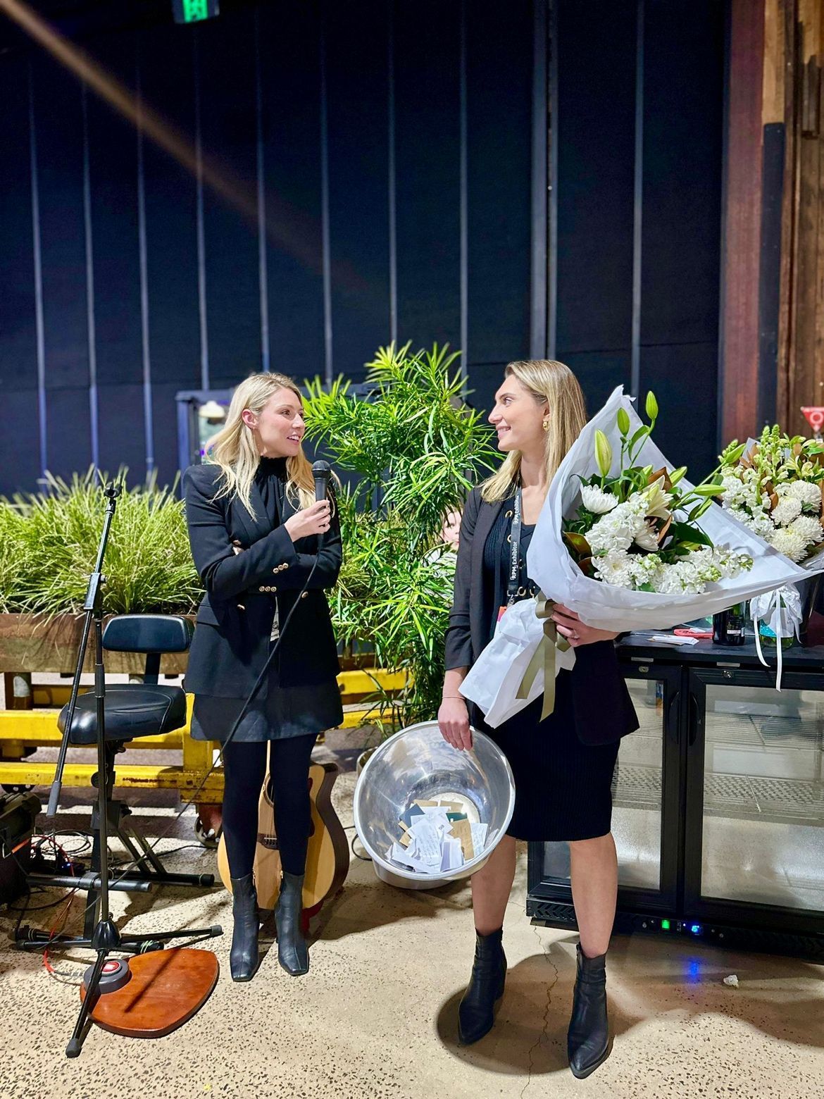 Two individuals stand on a stage, one holding a microphone and the other holding a large bouquet of white flowers.
