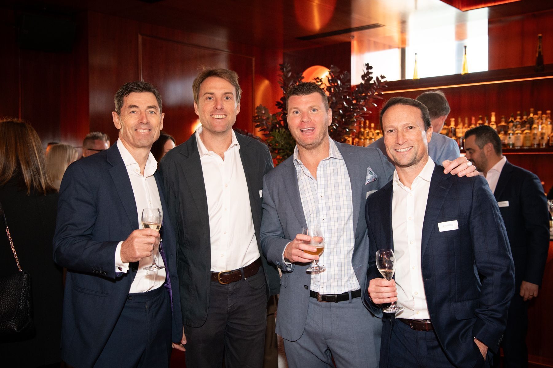 Four men in business attire smile while holding drinks at an evening event in a warm-toned, wood-paneled bar.