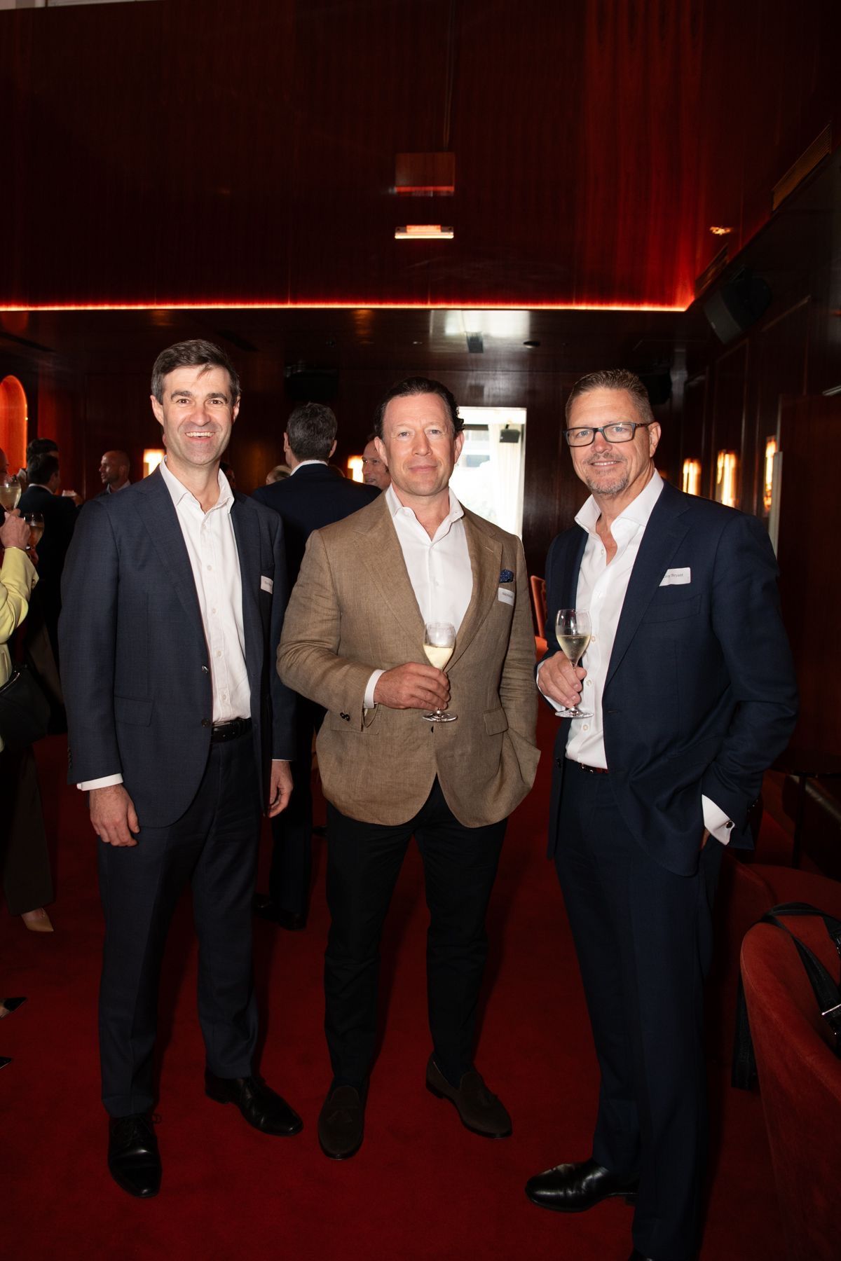 Three men in business attire stand smiling together in a warmly lit, red-carpeted interior setting.