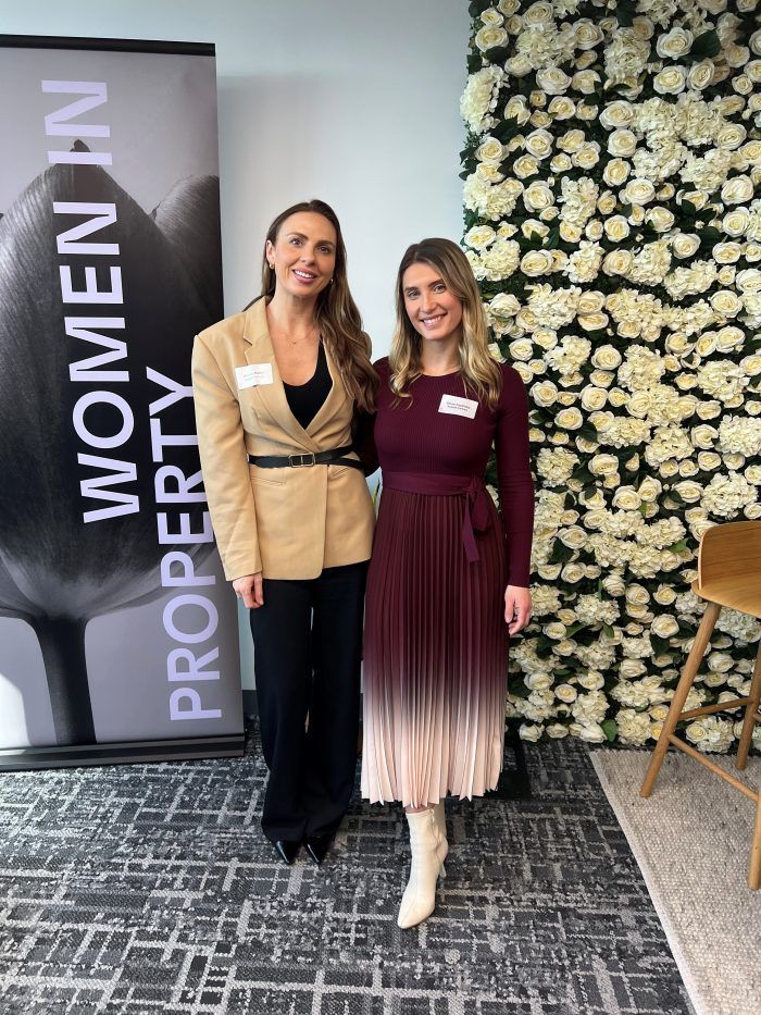 Two women stand smiling next to a Women in Property banner and a floral wall at a professional event.