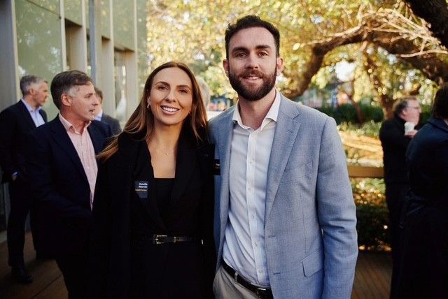 A woman and a man standing side-by-side smiling at an outdoor event, with other people in business attire in the background.