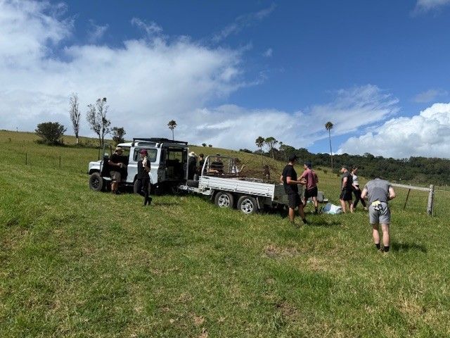 A group of people standing around a white utility vehicle and trailer in a grassy field under a sunny, blue sky.