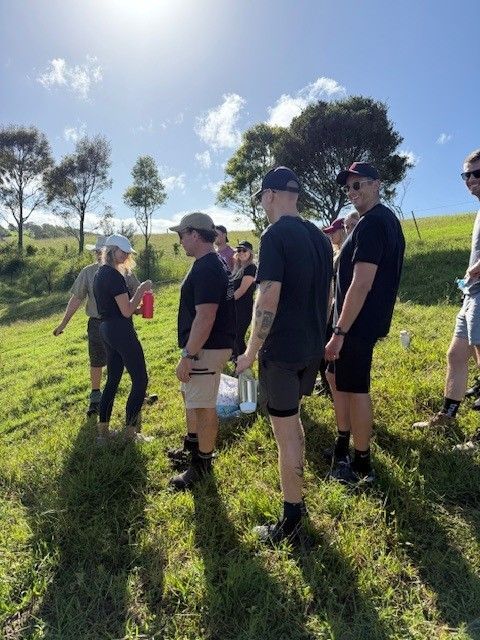 A group of people standing on a grassy hill outdoors on a sunny day, holding water bottles and chatting.