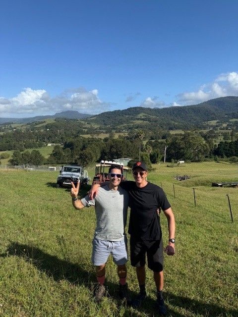 Two people stand in a sunny, grassy field with mountains in the background, one making a peace sign near a utility vehicle.