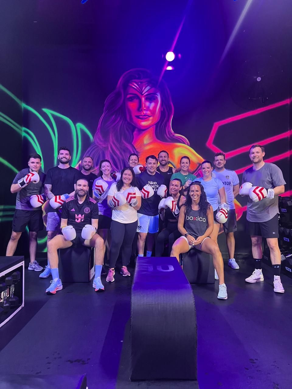 A group of people wearing boxing gloves pose for a photo in a gym with neon lighting and a large mural in the background.