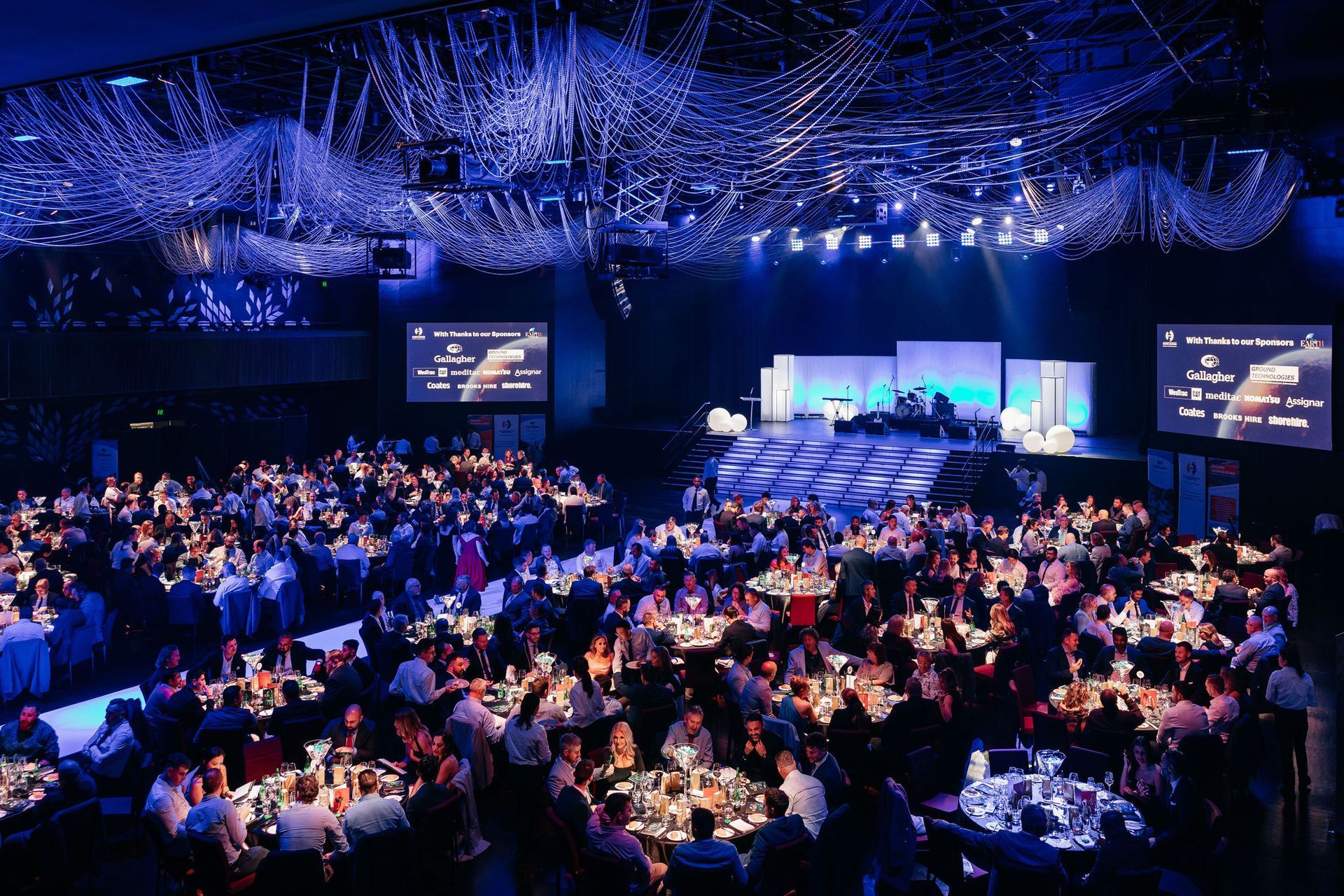 A formal gala dinner in a large, dimly lit hall with blue ceiling lights, round tables, and a stage with screens.
