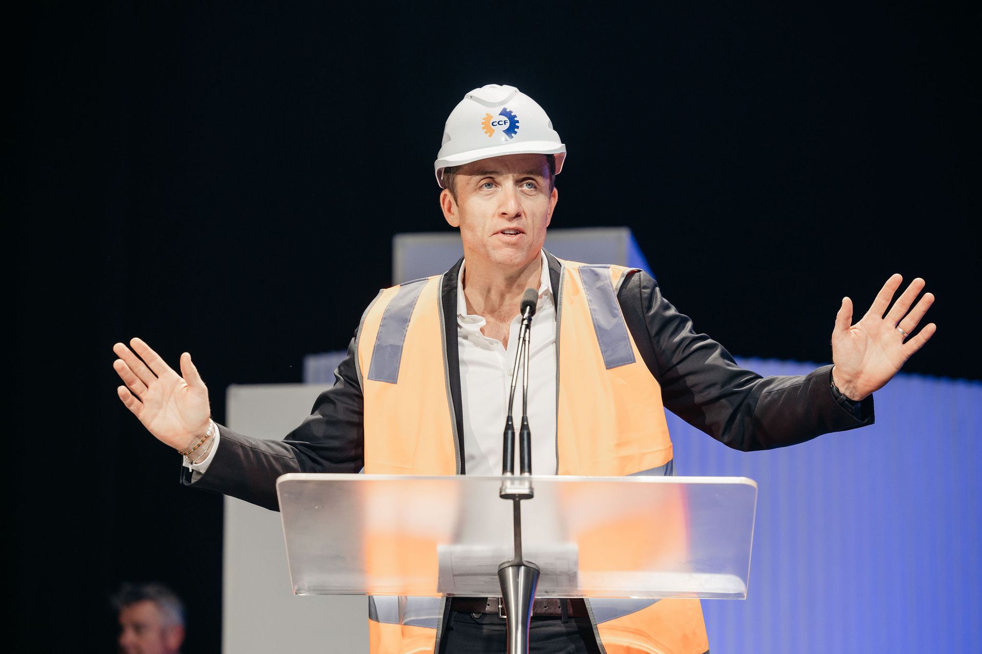 A person in a hard hat and high-visibility vest speaks at a podium with arms outstretched against a blue-lit backdrop.