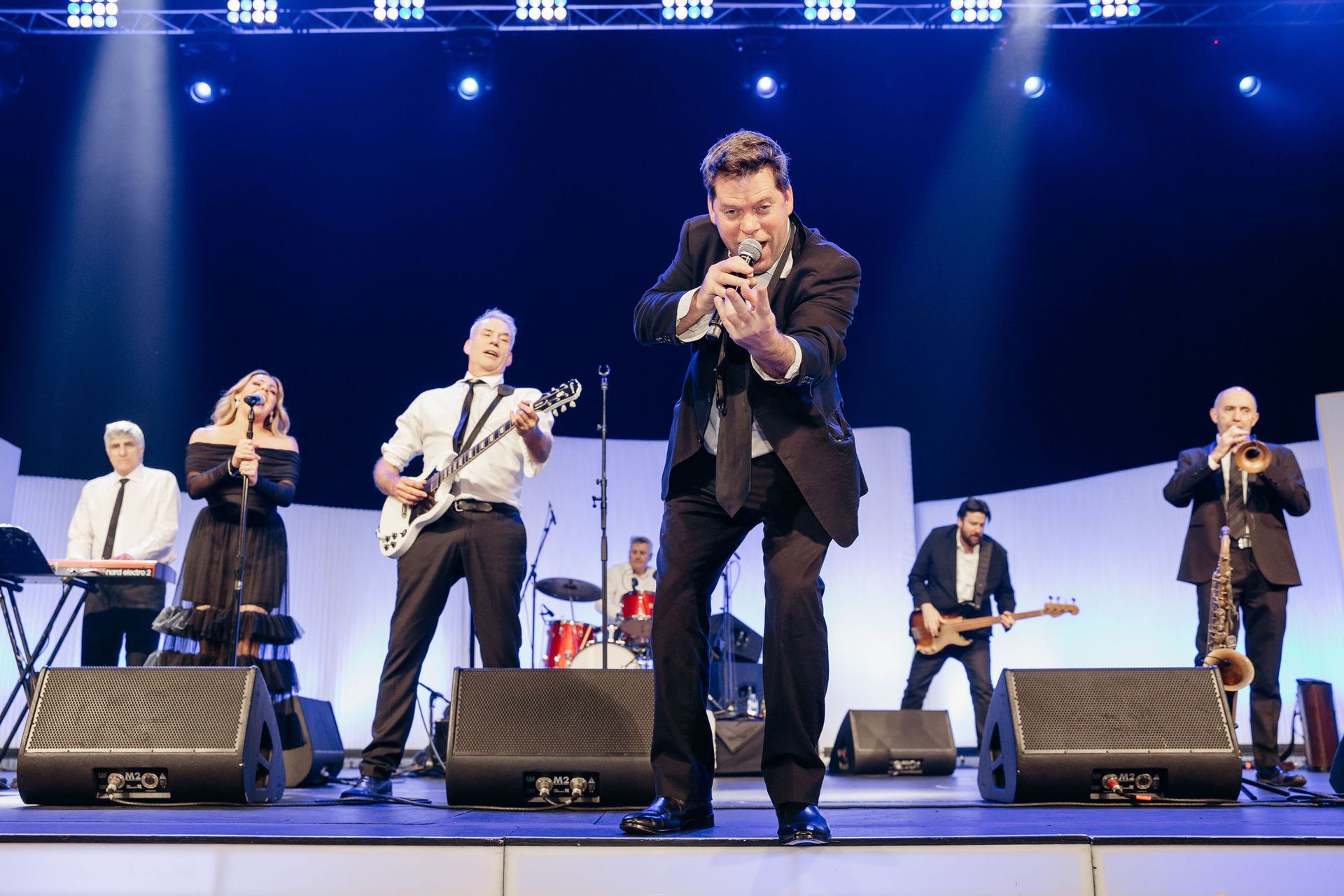 A musical band performs on stage under bright lights, with a lead singer at the center holding a microphone.