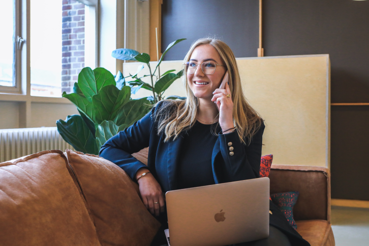 A smiling person in a dark blazer sits on a leather couch with a laptop, speaking on a phone in a modern office space.