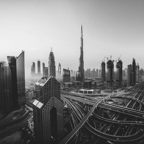 Black and white aerial view of the Dubai skyline with the Burj Khalifa and a large highway interchange at dusk.