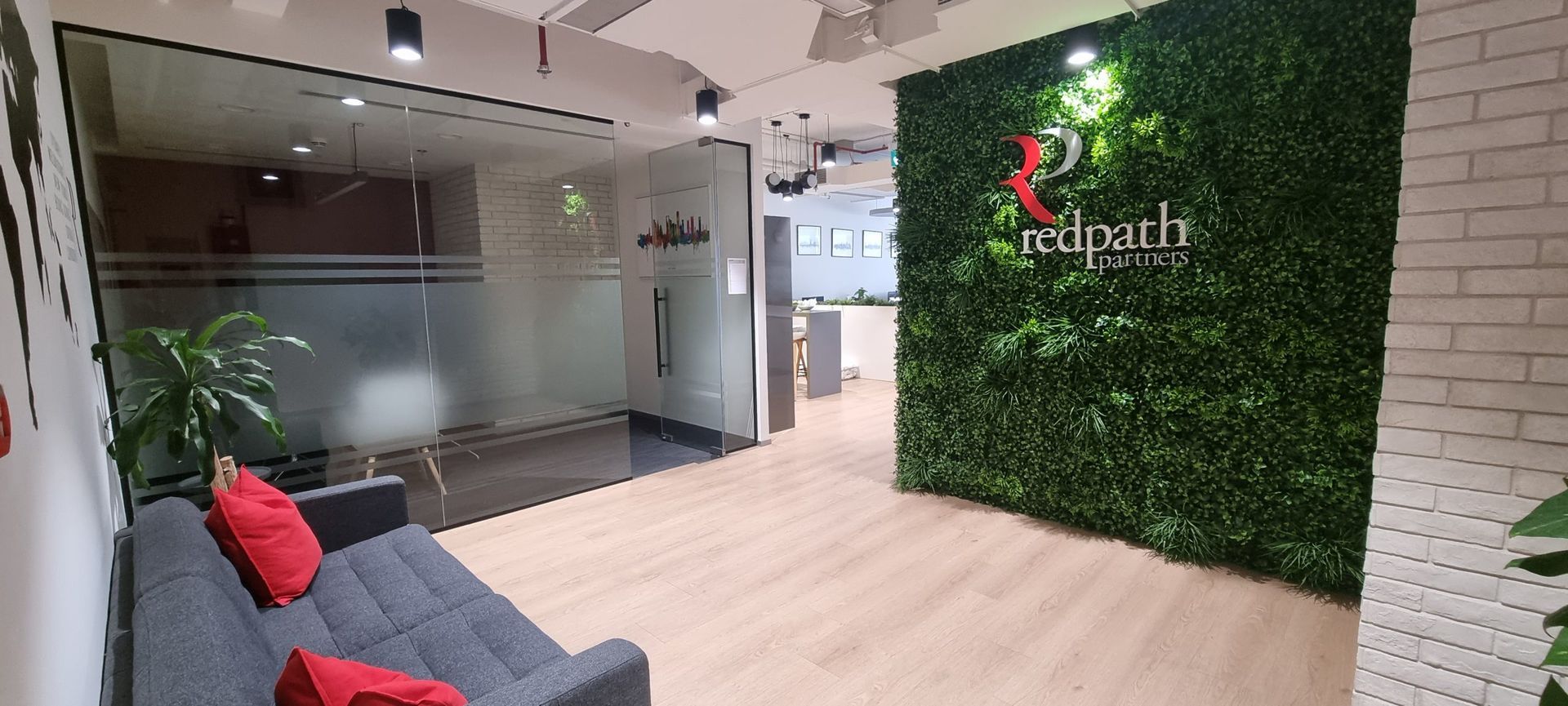 Modern office lobby with light wood floors, a grey sofa with red pillows, and a prominent green plant wall with a logo.