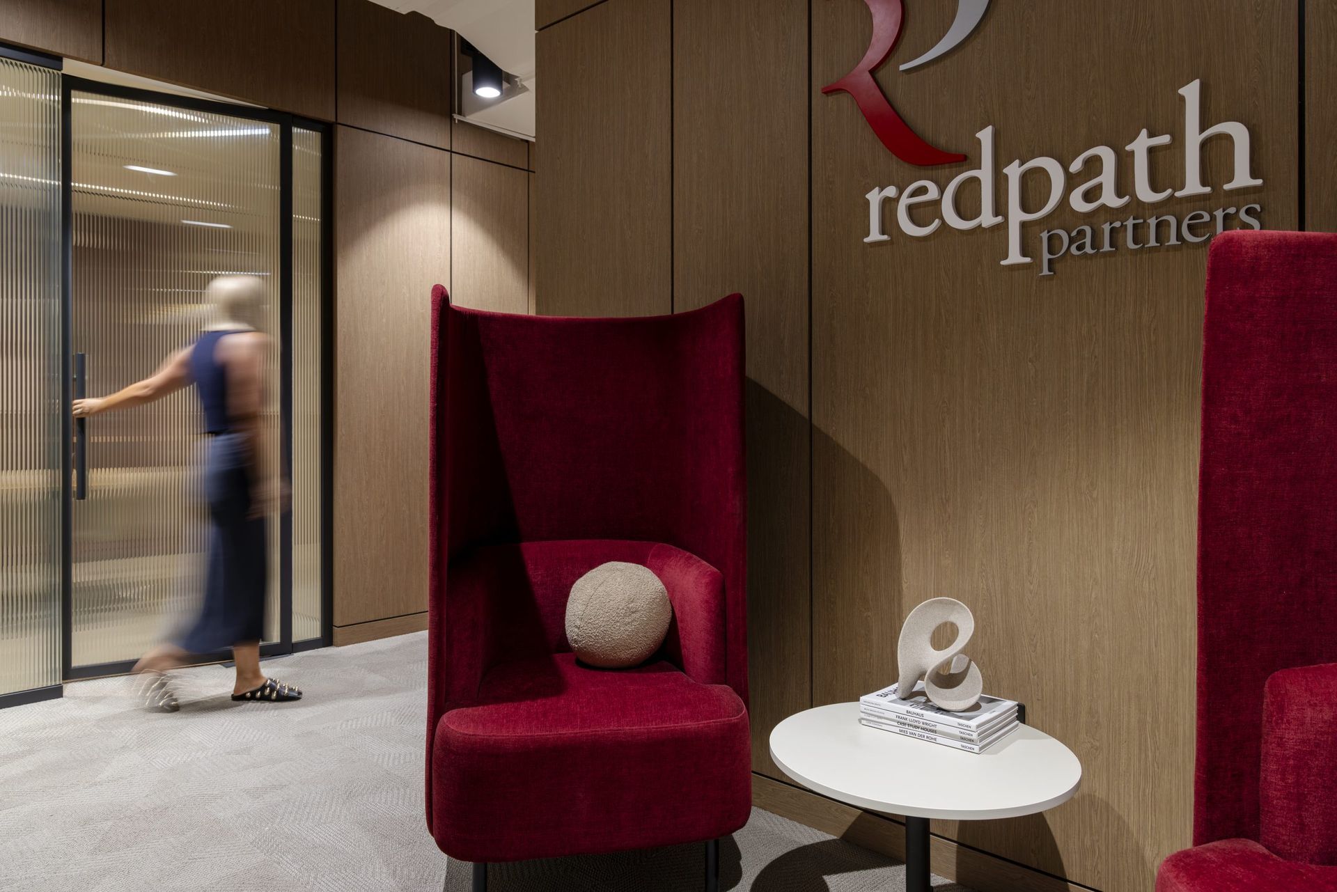 A person walks past a high-backed red armchair and a small table with a sculpture in a wood-paneled office lobby.