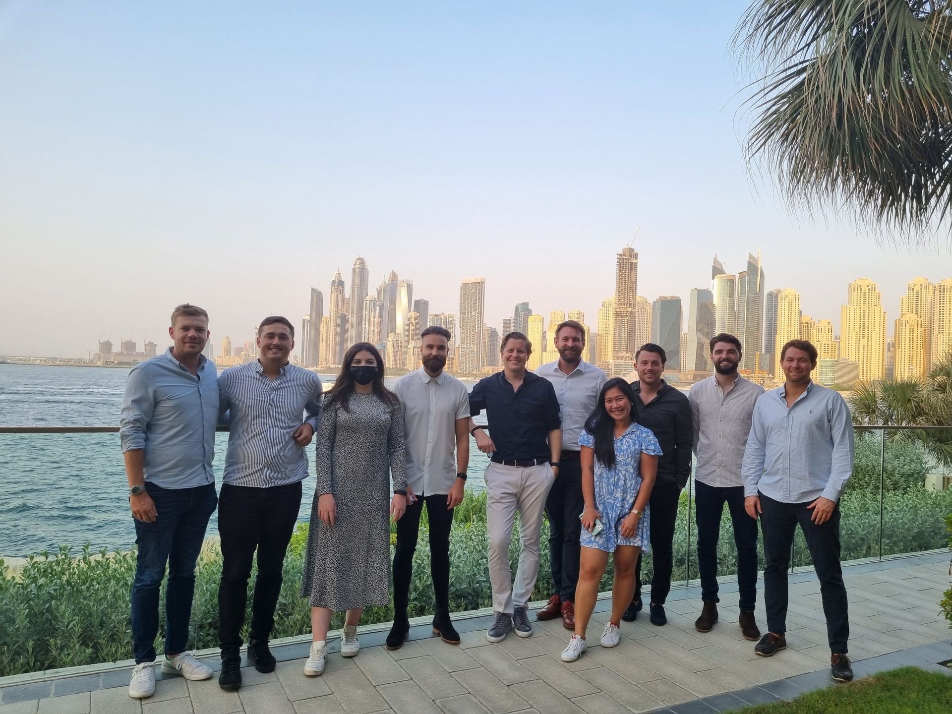 A group of eleven people standing in a row on a waterfront promenade, posed before the Dubai Marina skyline at dusk.