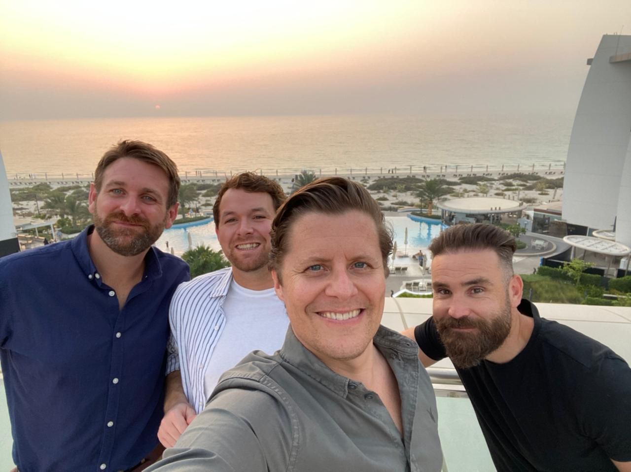 Four men smiling for a selfie on a balcony overlooking a coastal resort at sunset.