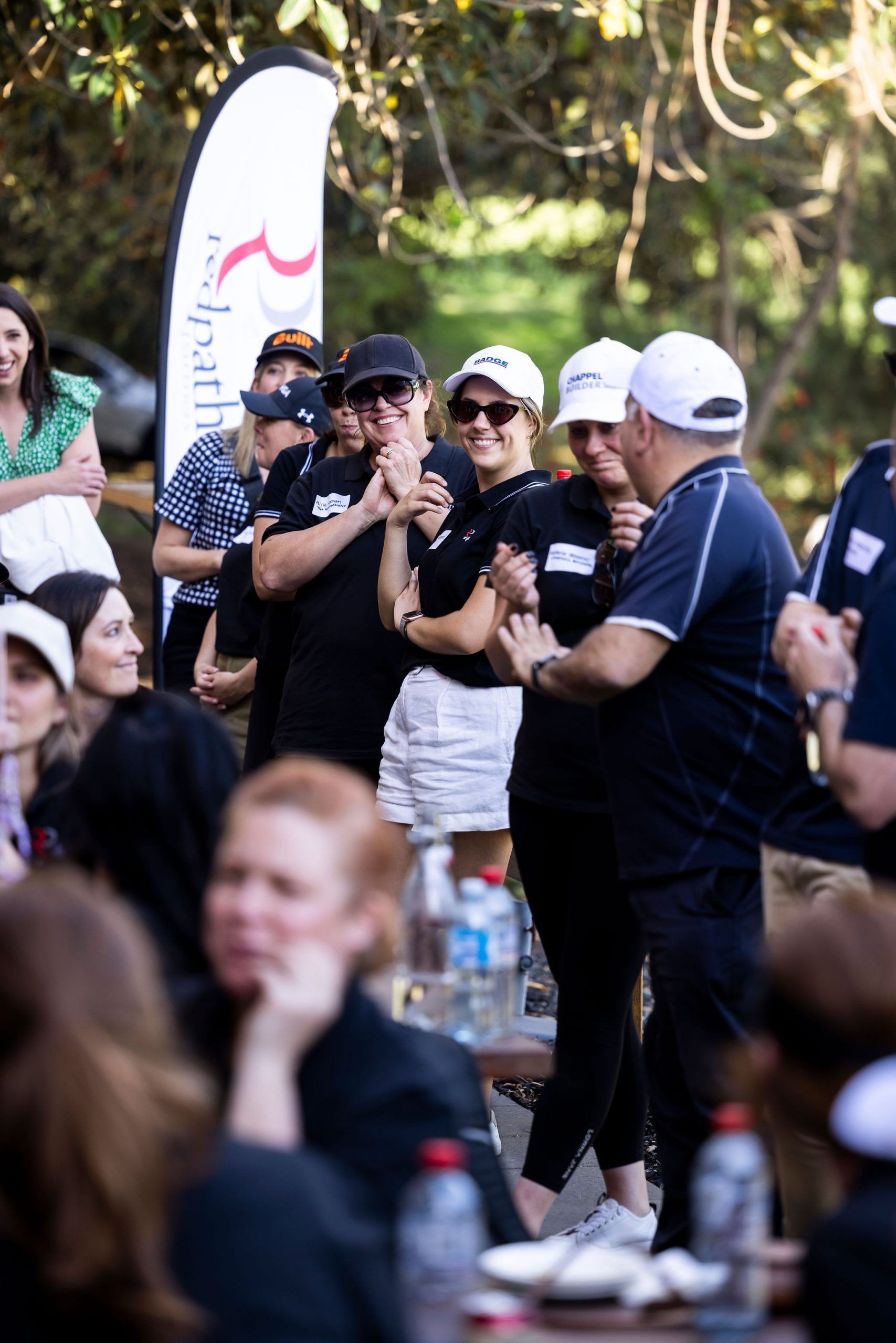 People gathered outdoors at a social event, with one person speaking into a microphone near a banner.