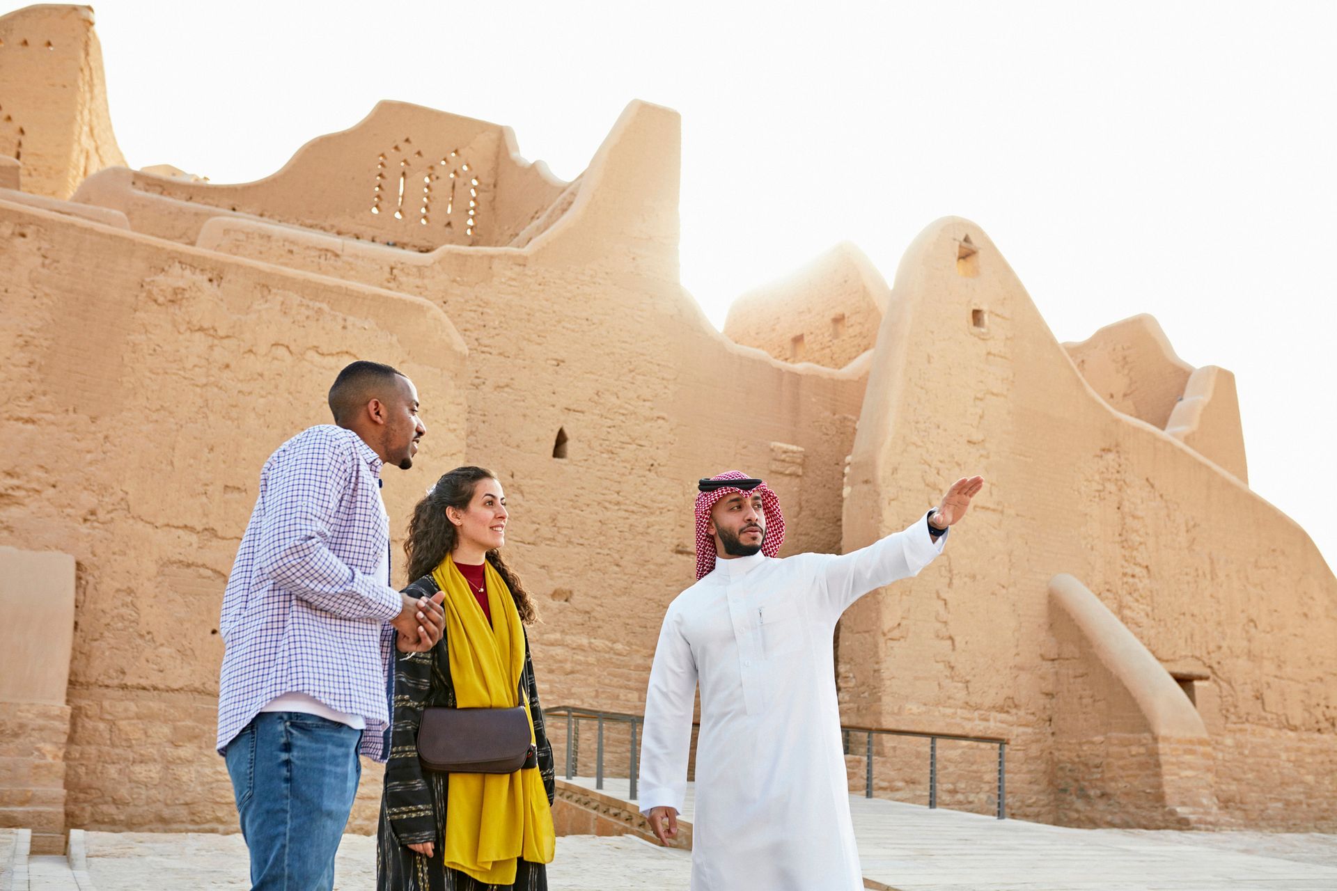 A guide points toward the historic mud-brick walls of At-Turaif as two visitors listen intently.