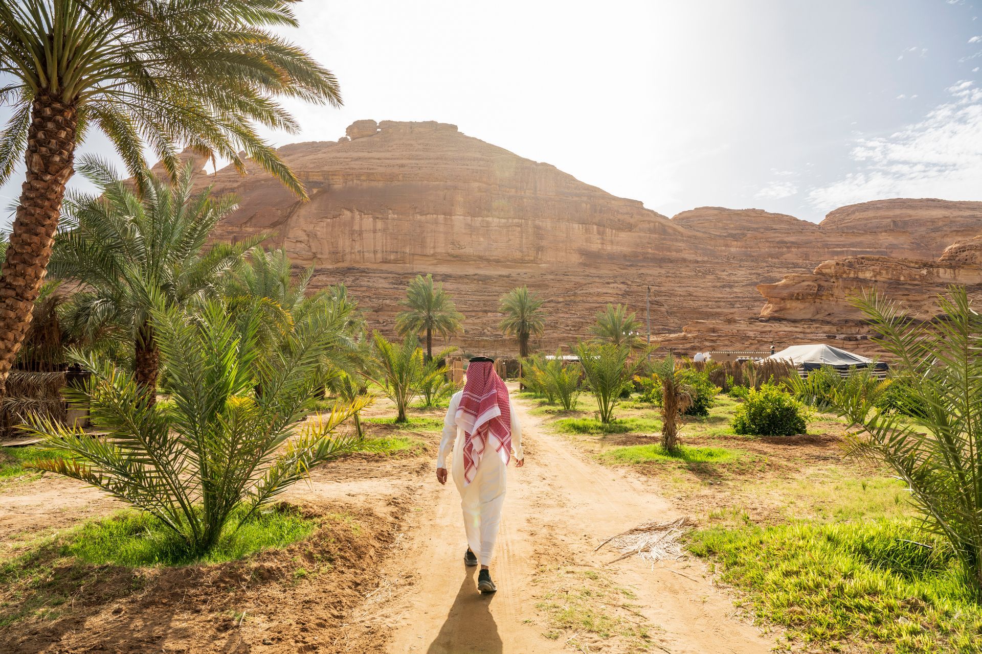 A person in traditional white attire walks along a dirt path through a sunlit palm grove beneath a rocky desert cliff.