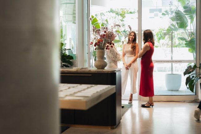 Three people stand in a bright, modern interior by a large window, engaging in conversation near a desk with a flower vase.