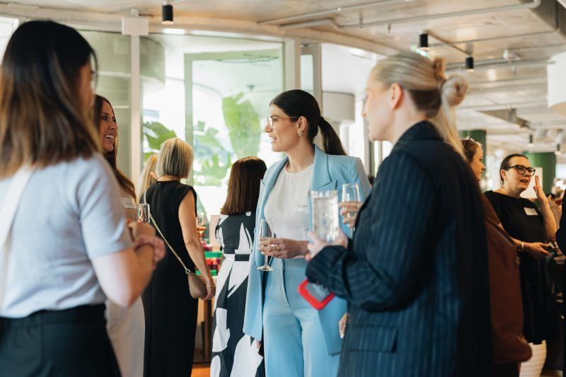 A group of people networking at an indoor professional event, engaged in conversation while holding drinks.
