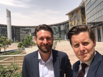 Two men in suits pose for a selfie in front of a modern, curved glass building and a tall tower under a sunny sky.