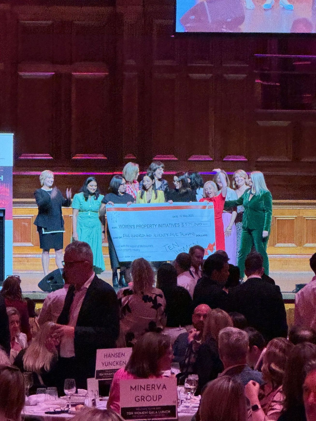A group on stage holds a large, oversized check during an indoor event in a wood-paneled hall with audience seating.