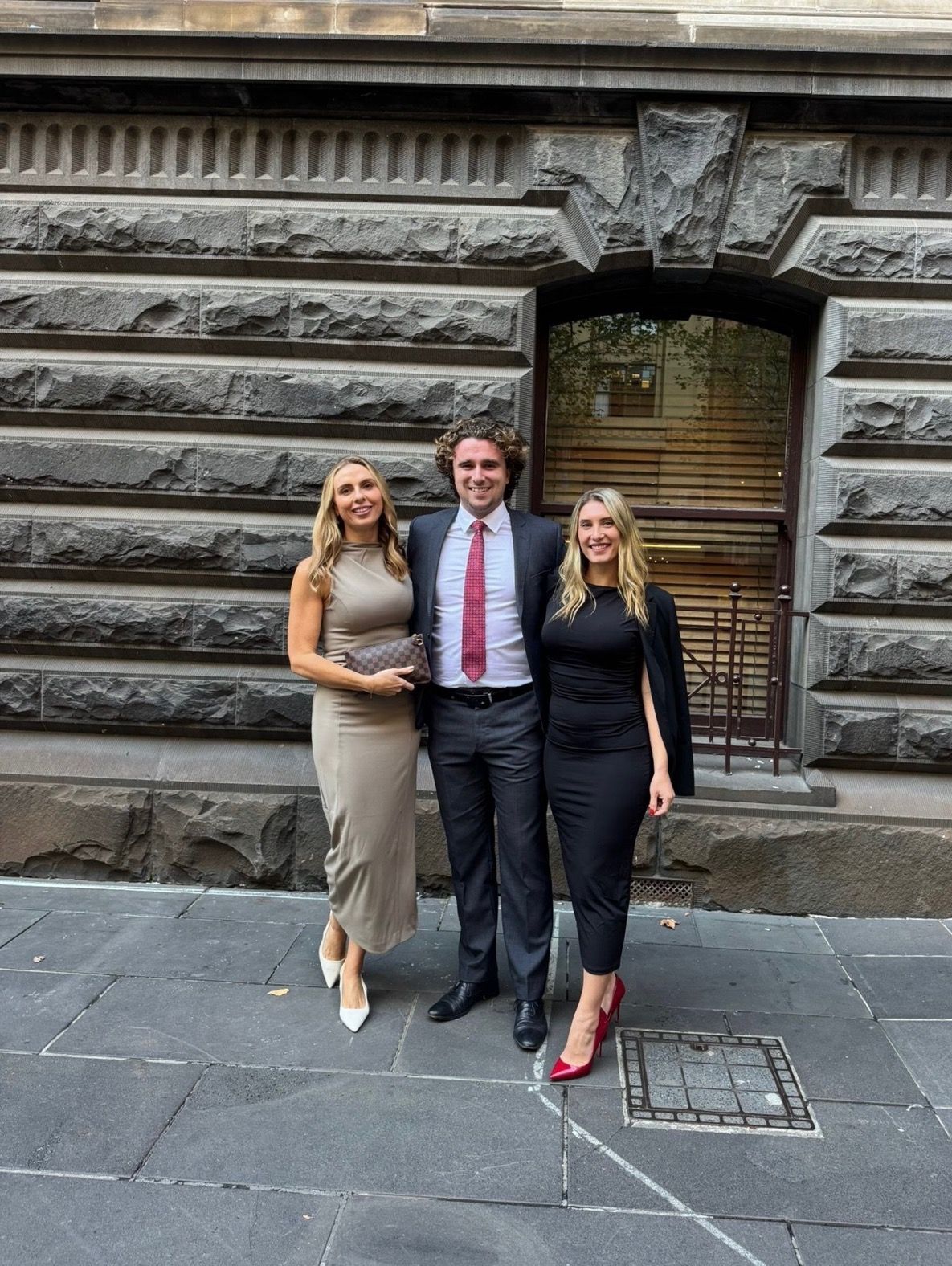 Three people stand smiling on a stone sidewalk in front of a textured gray masonry building.