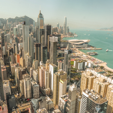 High-angle view of Hong Kong's dense urban skyline, featuring numerous skyscrapers, the harbor, and a waterfront building.
