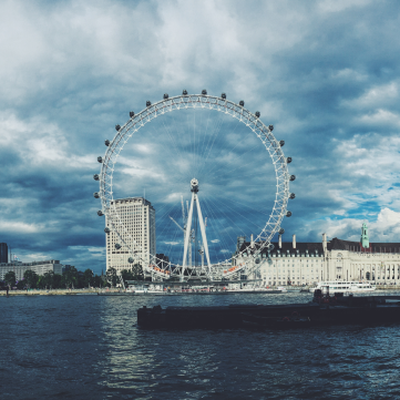 The London Eye observation wheel stands by the River Thames against a backdrop of dark, cloudy skies and city buildings.