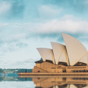 The iconic, sail-like white roofs of the Sydney Opera House stand against a cloudy sky above calm, reflective water.