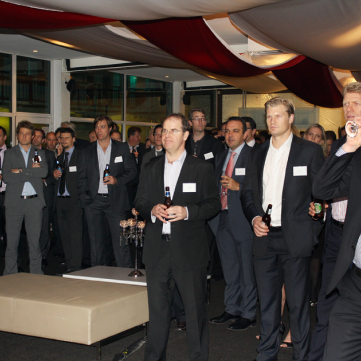 A group of men in business attire holding drinks, standing in a decorated room for an evening social gathering.