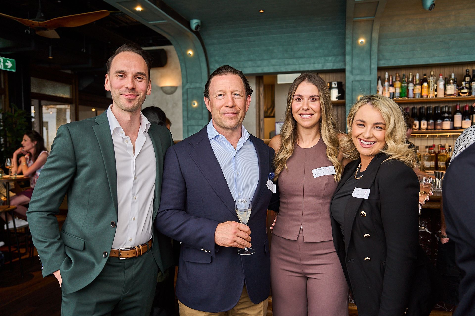 Four people stand side-by-side in a bar setting, smiling and posing for a photo while holding drinks.