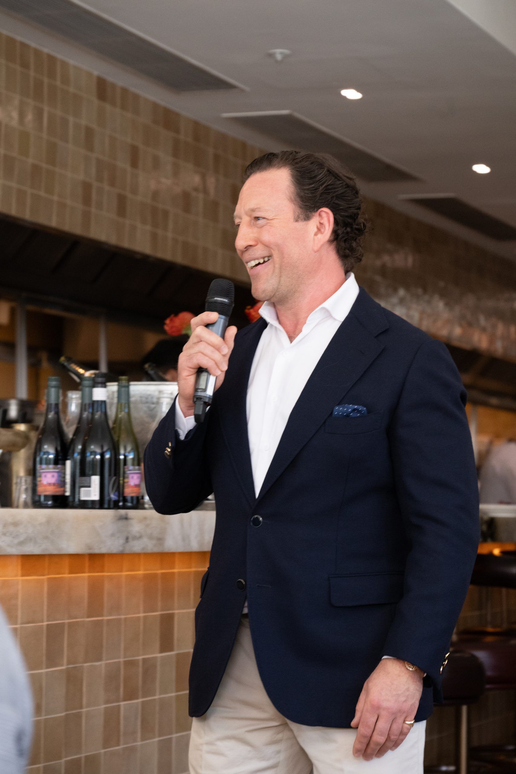 A smiling person in a dark blazer and white shirt speaks into a microphone in a restaurant setting.