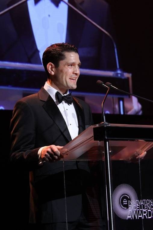 A person in a black tuxedo and bow tie speaks at a podium during the Innovation Excellence Awards ceremony.