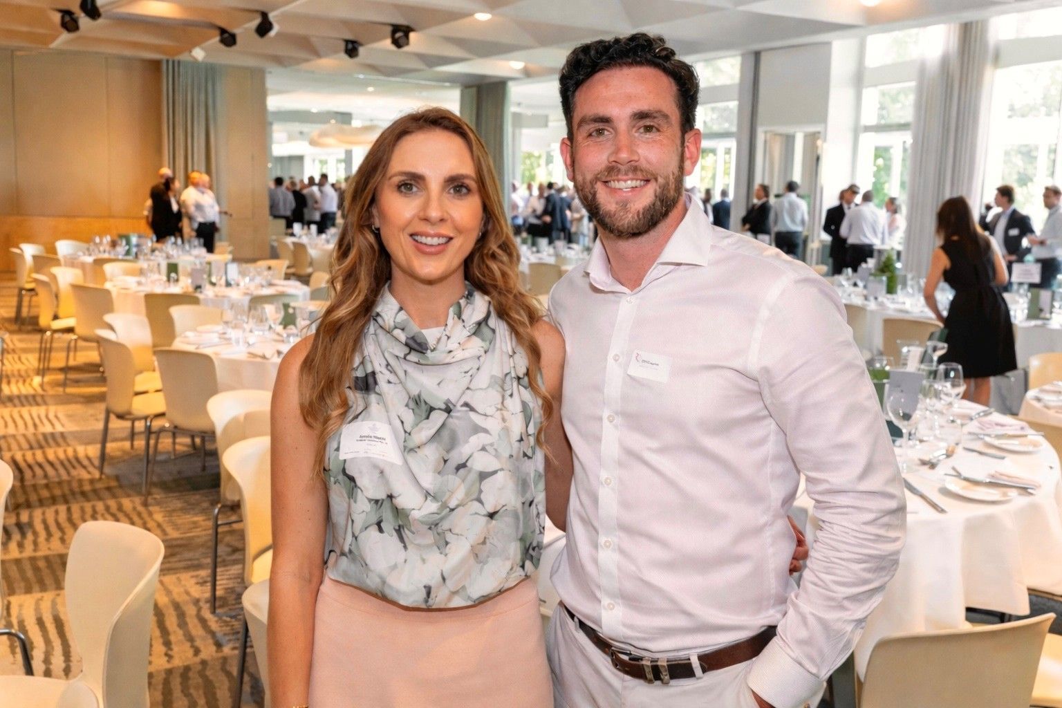 A man and woman standing together in a formal event space with tables and guests in the background.
