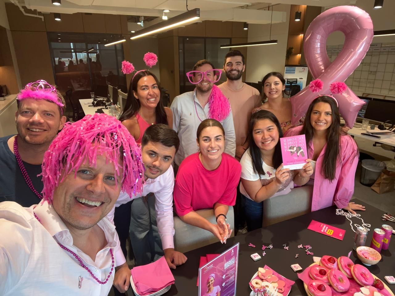 A group of colleagues wearing pink accessories in an office for a breast cancer awareness event with donuts on a table.