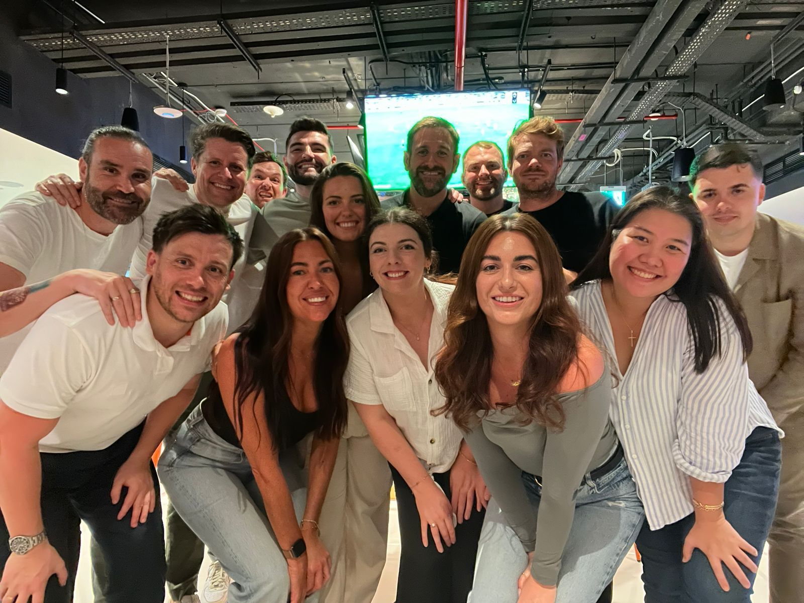 A group of people smiling and posing for a photo indoors, with a television screen in the background.