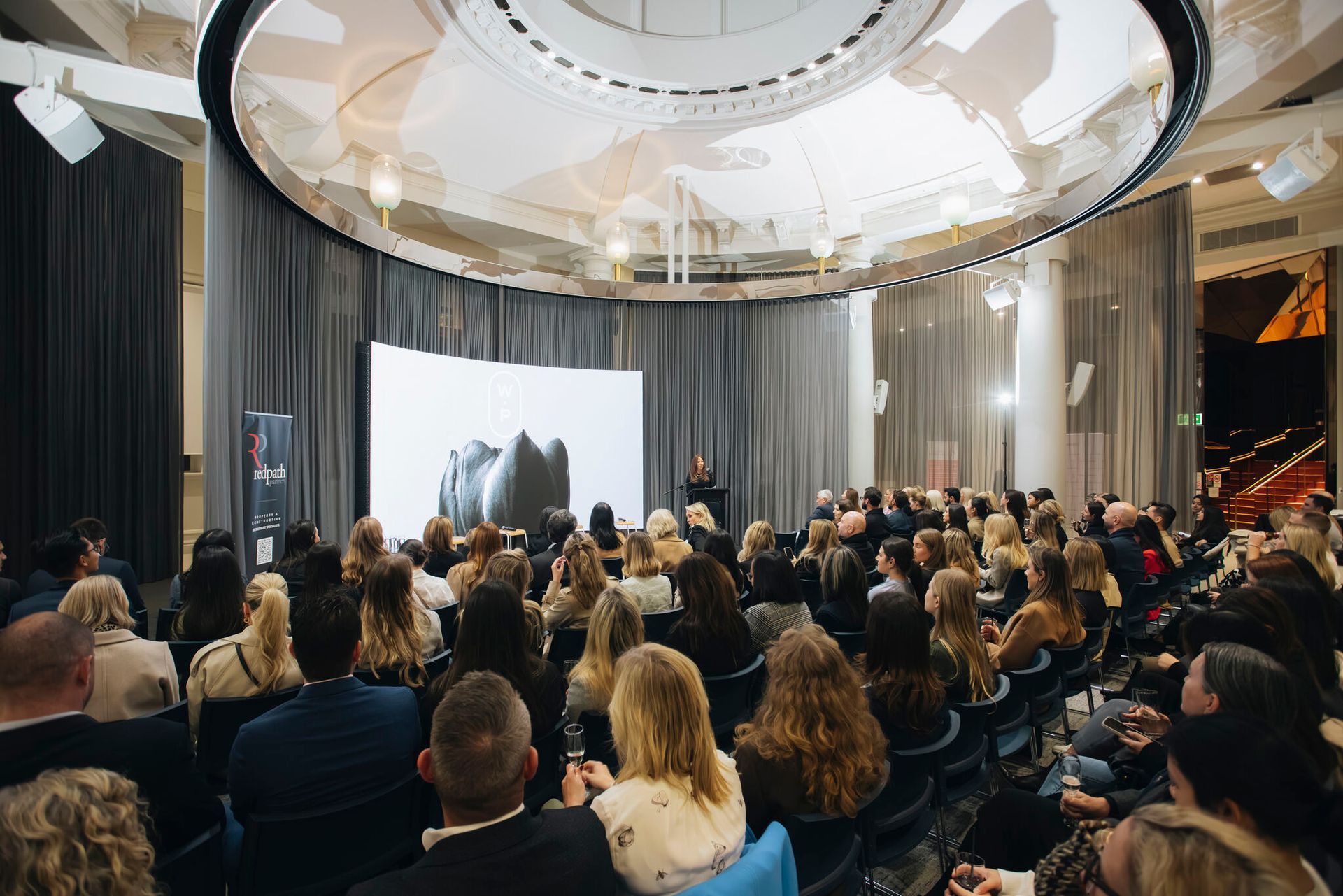 A large audience seated in a semi-circular, modern event space watching a presentation on a curved screen.