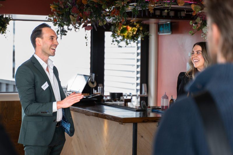 A person in a suit smiles while speaking to a group in a decorated bar area.
