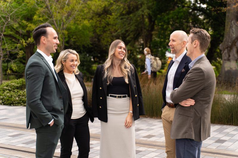 A group of four professionals in business casual attire stand on a paved pathway, talking and smiling in a park.