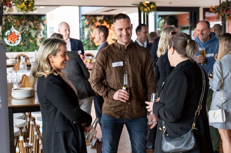 People socialize at an indoor event, talking and holding drinks in a bright, decorated space.