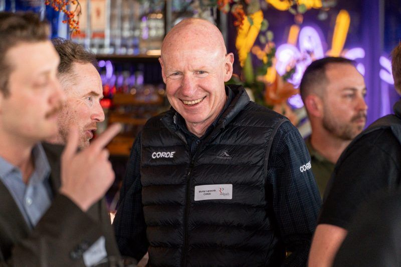 A group of people at a social gathering, with a smiling individual in a black puffer vest prominently in the foreground.