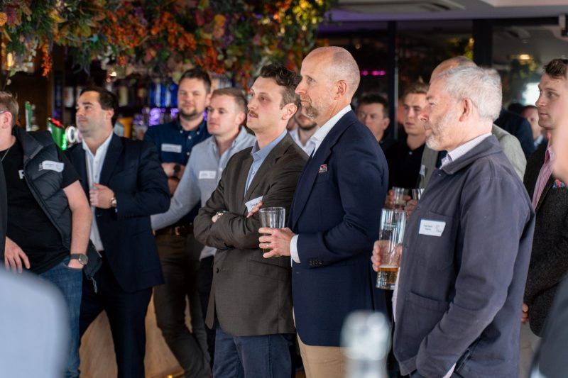 A group of people in business casual attire standing in a room with a decorative ceiling, listening to a presentation.
