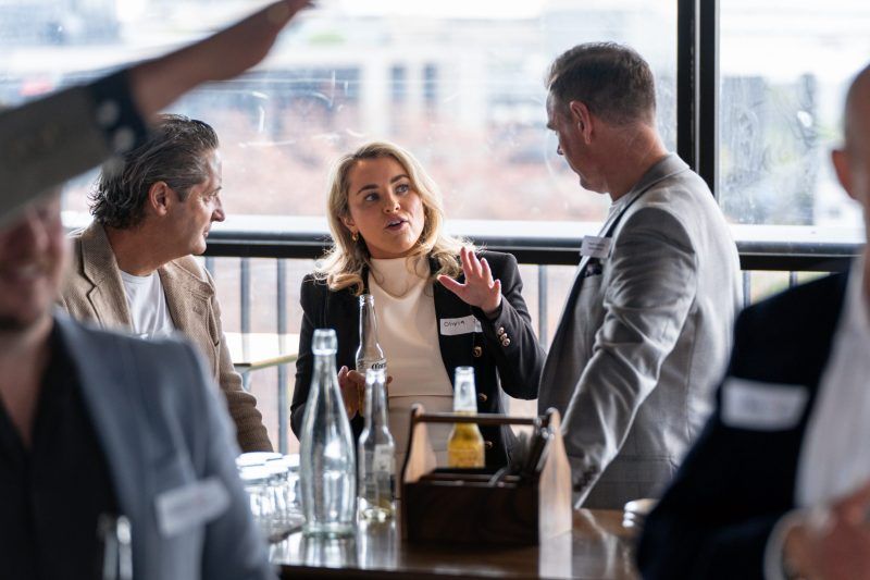 A group of people wearing name tags talk and gesture while standing around a high table in a bright, modern indoor space.