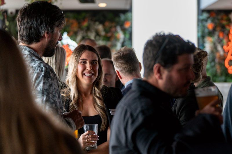 People socialize in a warmly lit, indoor space with foliage, holding drinks and smiling at a networking event.