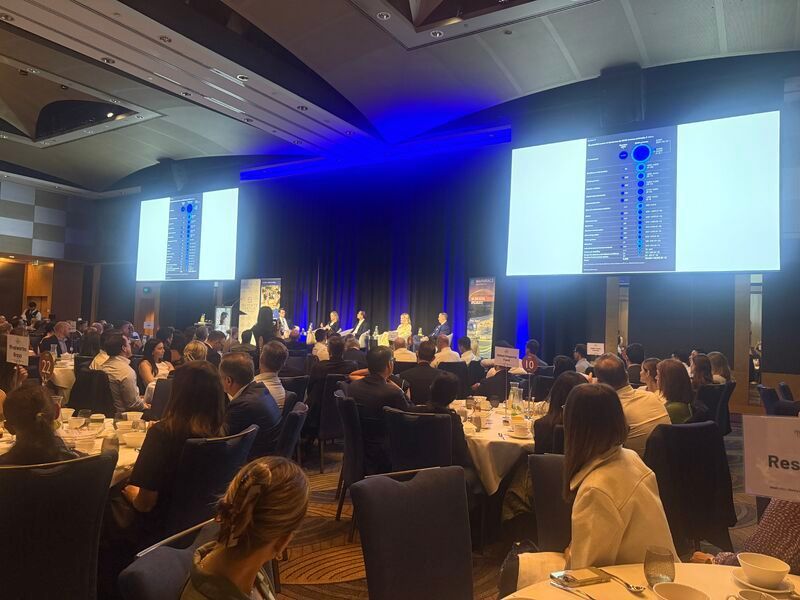 A large audience sits at round tables in a dim hotel ballroom facing a stage with two large screens showing data charts.