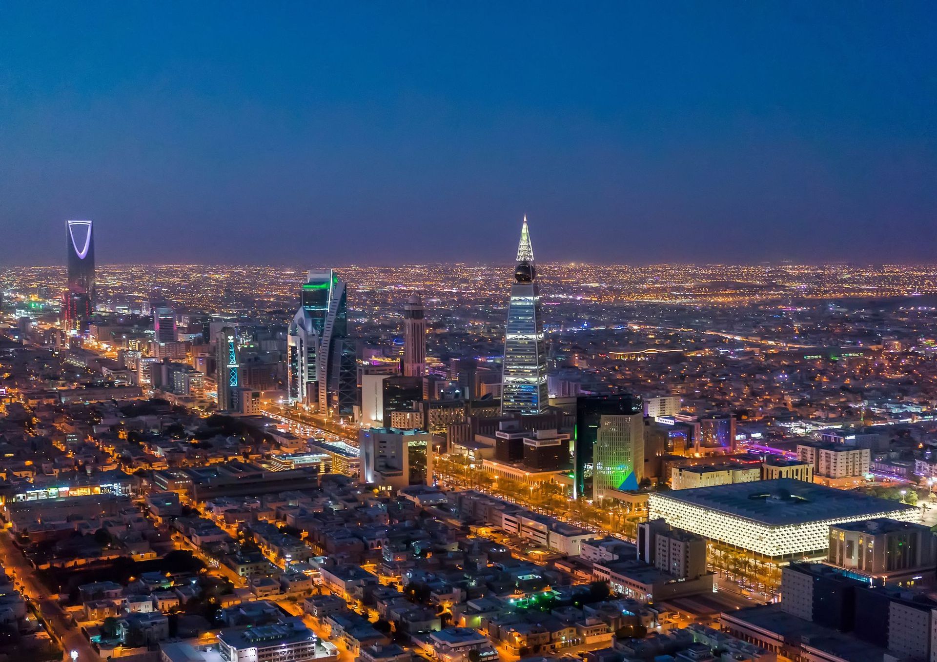 A wide-angle aerial night view of the illuminated Riyadh city skyline, featuring modern skyscrapers and grid-like streets.