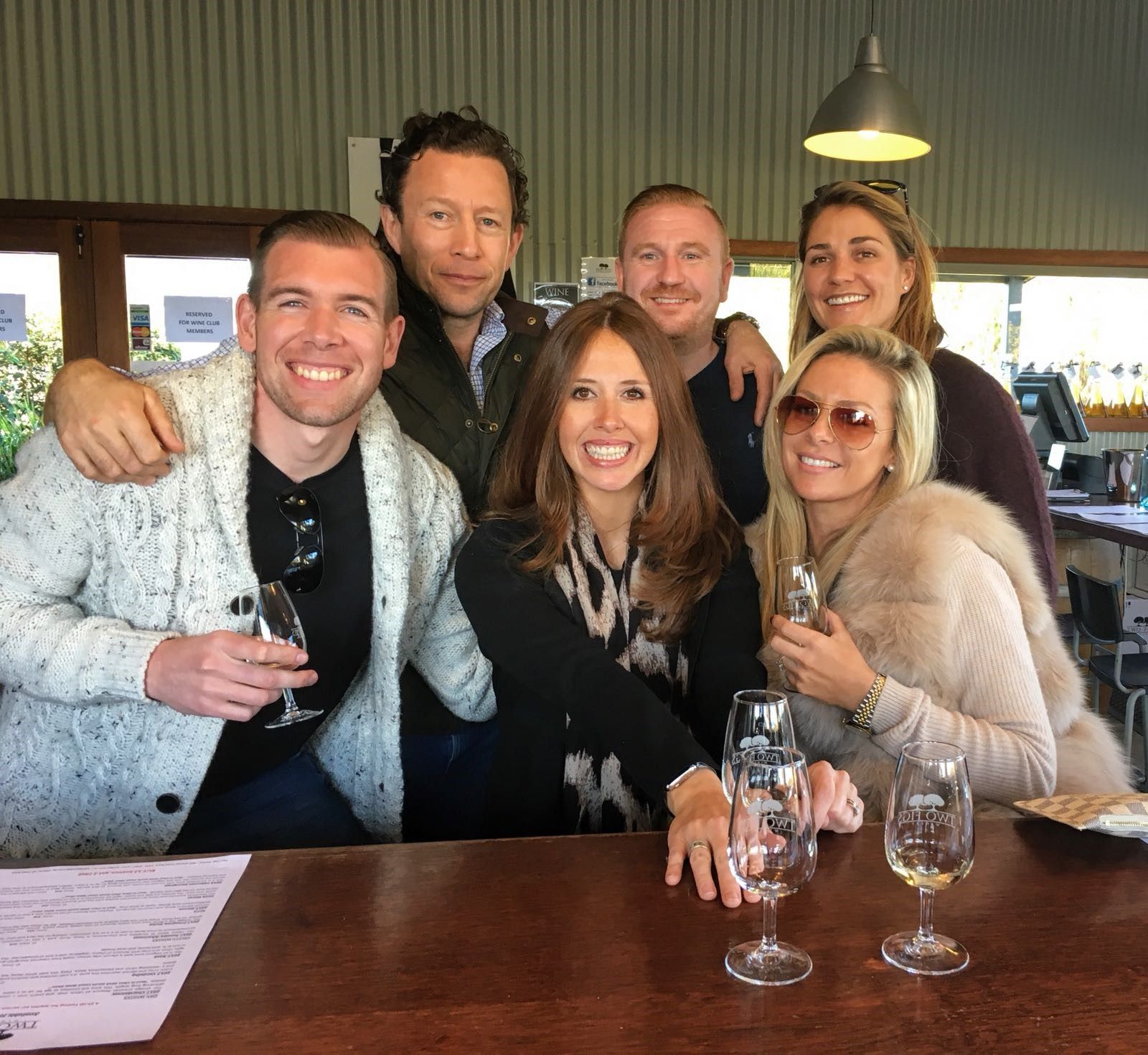 Six people smiling while standing together at a wooden bar with glasses of wine in an indoor setting.