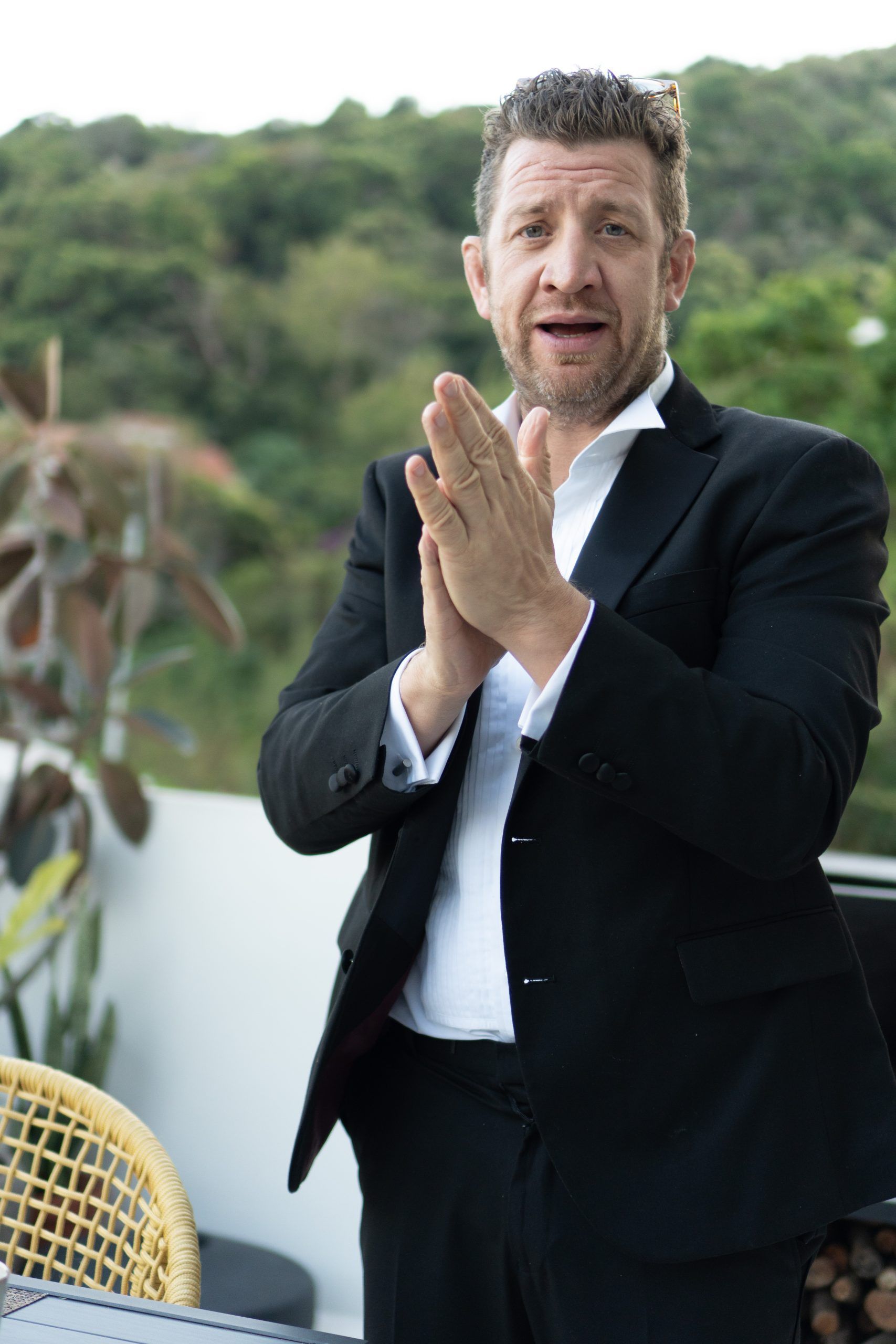 A person in a black suit and white shirt stands outdoors with hands pressed together, looking forward on a hillside.