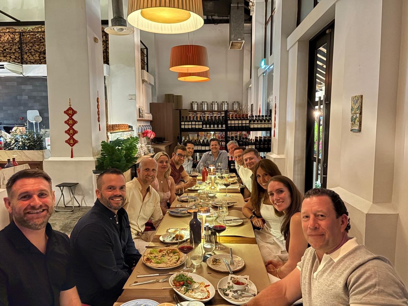 A group of people sitting at a long table in a well-lit restaurant, eating and talking during a social dinner.